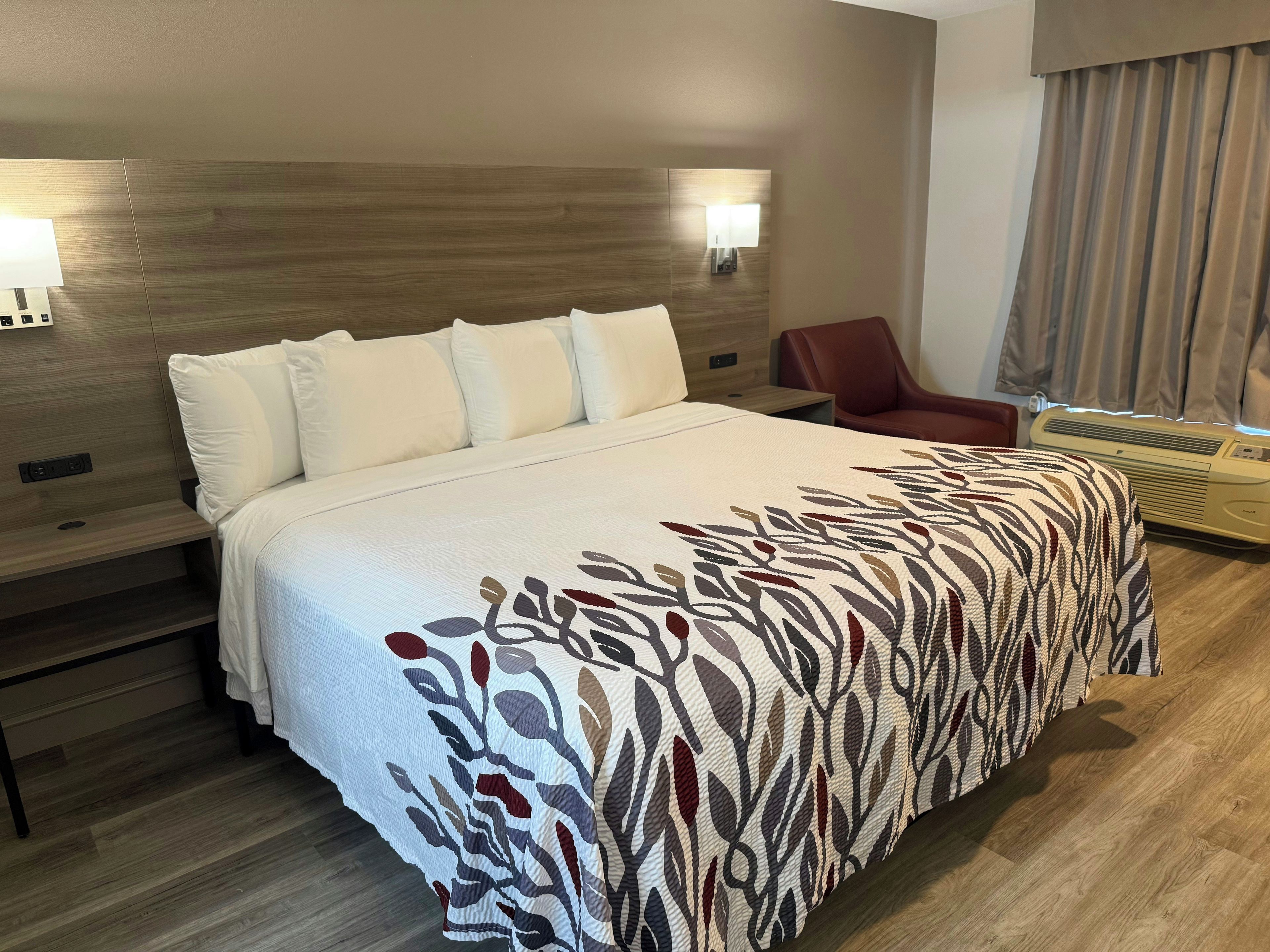 A hotel room with a large bed, two wall-mounted lights, a red chair, and a window with curtains.
