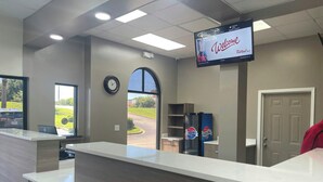 A reception desk with a digital screen displaying 'Welcome' and 'Red Roof Inn.' There is a clock on the wall, a window with a view of a parking lot, and a refrigerator with 'Carolina's' branding. - Red Roof Inn Orangeburg (Orangeburg)