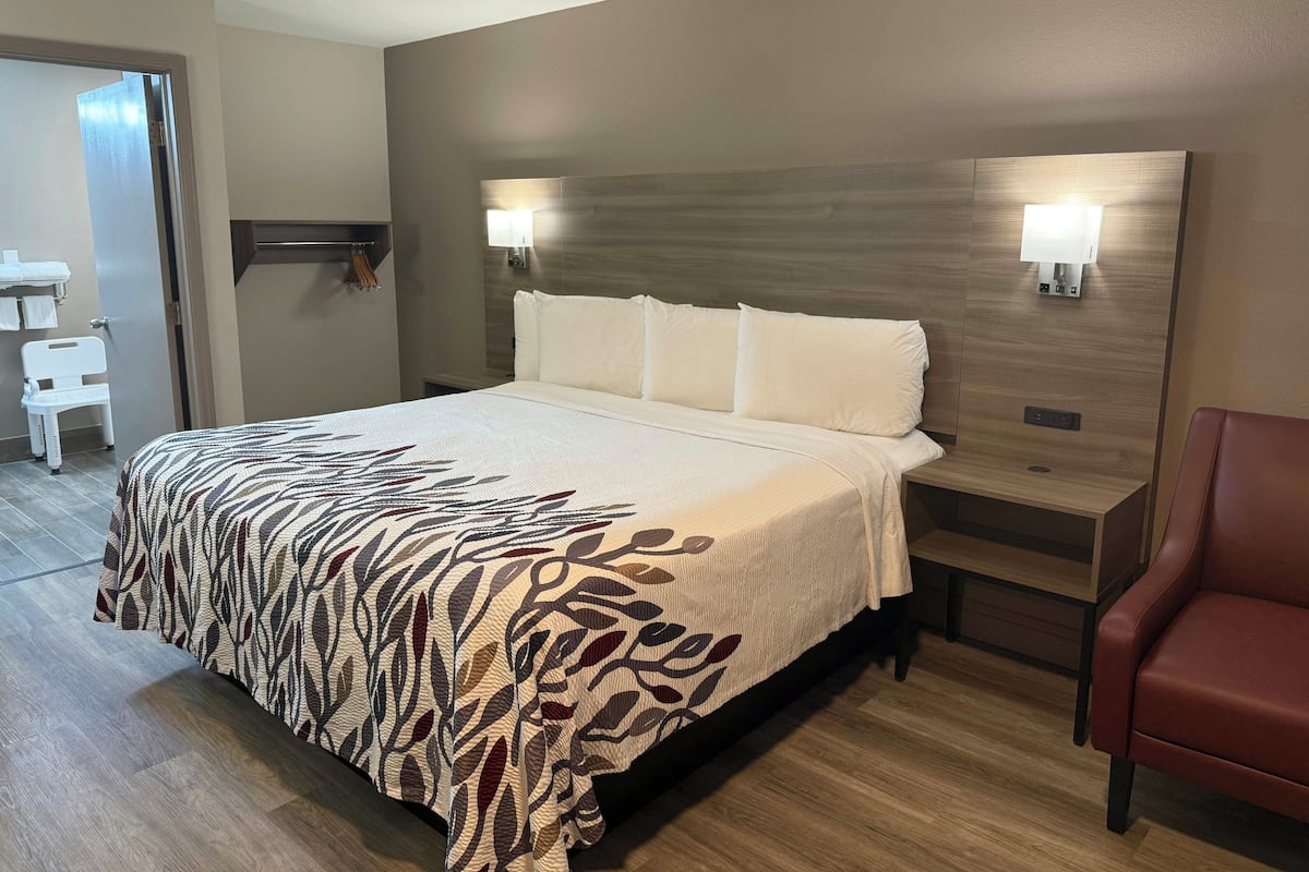 A hotel room with a bed, a nightstand, a chair, and a bathroom visible through an open door.