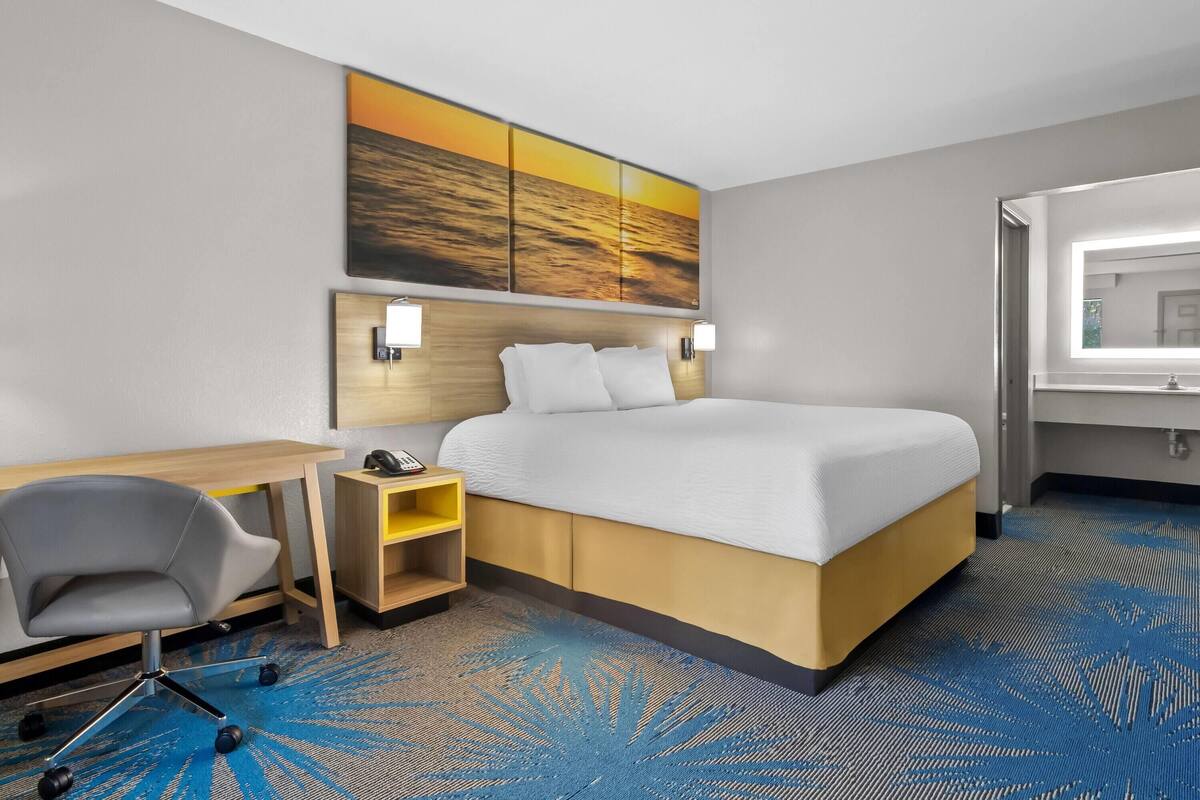 A hotel room with a bed, desk, chair, and a large painting of a sunset.