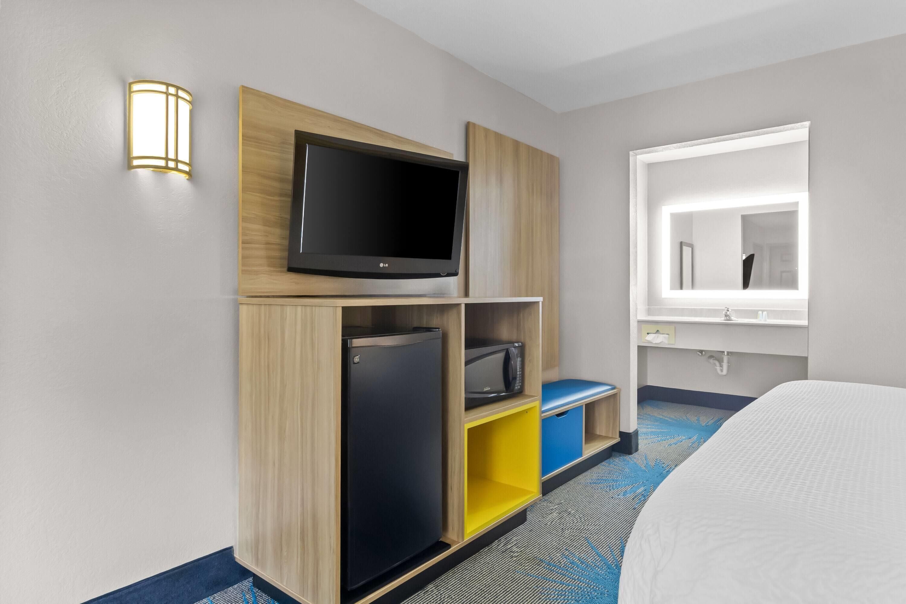 Ein Hotelzimmer mit Flachbildfernseher, einer Minikühlschrank, einer Mikrowelle, einem Bett, einem Fenster mit Spiegel und einer wandmontierten Leuchte.