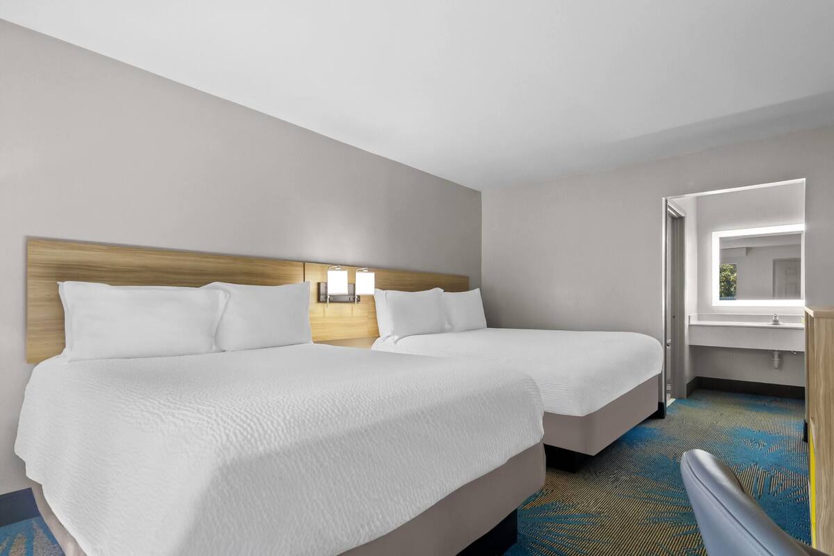 A hotel room with two beds, a wooden headboard, a nightstand with a lamp, a window, and a bathroom visible through an open door.