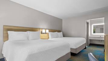 A hotel room with two beds, a wooden headboard, a nightstand with a lamp, a window, and a bathroom visible through an open door.