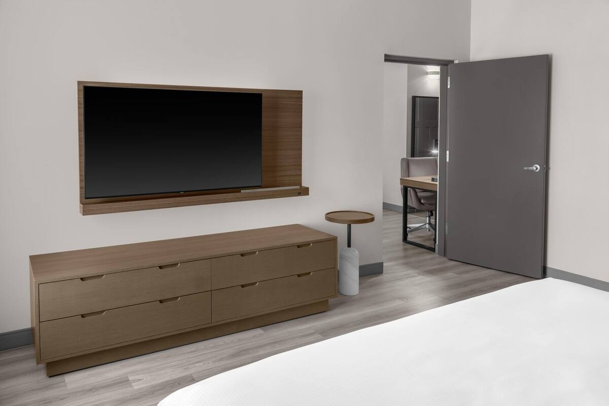 A modern bedroom with a large bed, a wooden dresser, a TV mounted on the wall, and a door leading to another room.