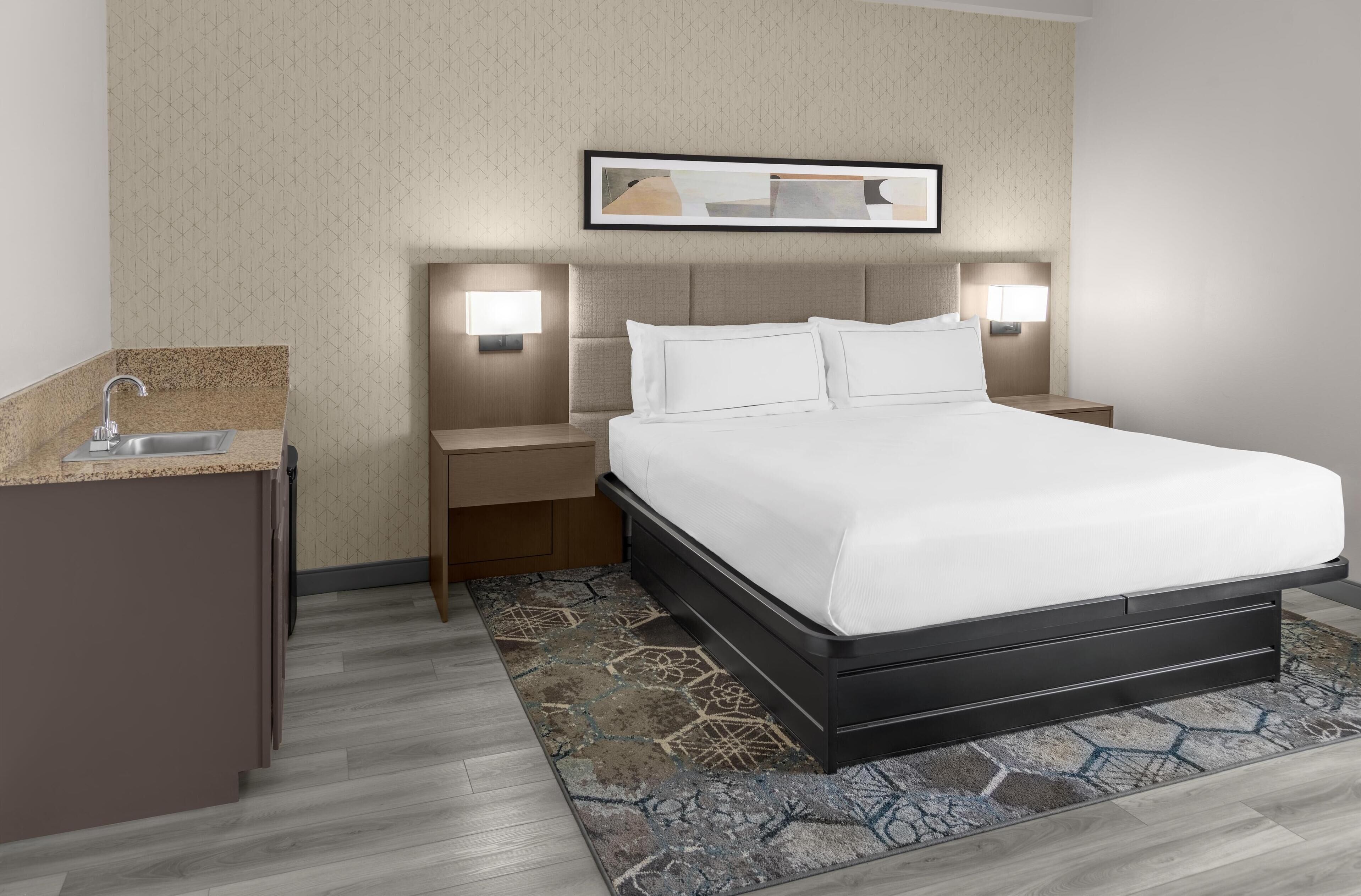 A hotel room with a bed, bedside tables, a nightstand, a sink, and a mirror.