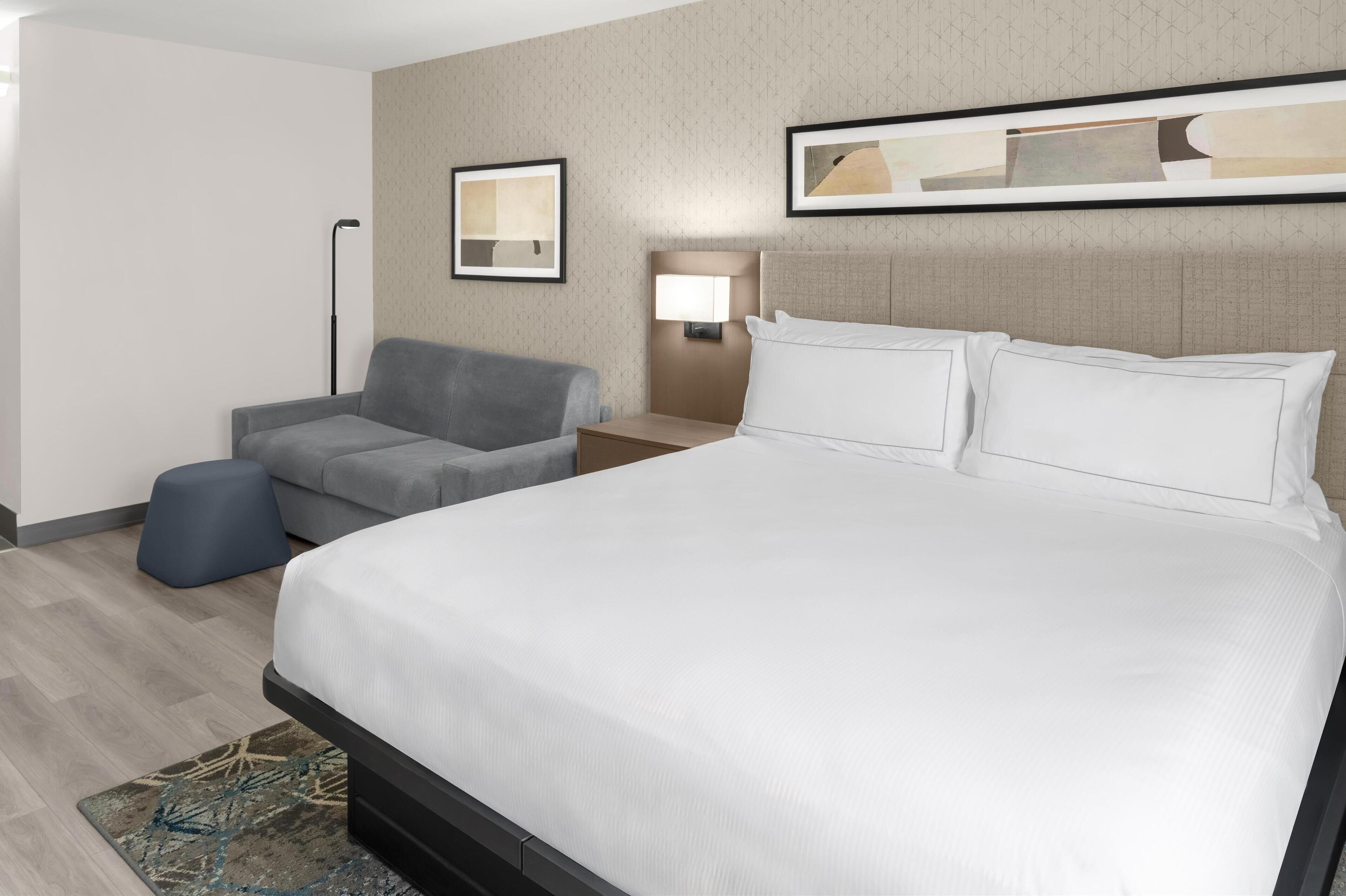Een hotelkamer met een groot bed, een grijze fauteuil, een klein poufje, een nachtkastje met een lamp, en een kunstwerk aan de muur.