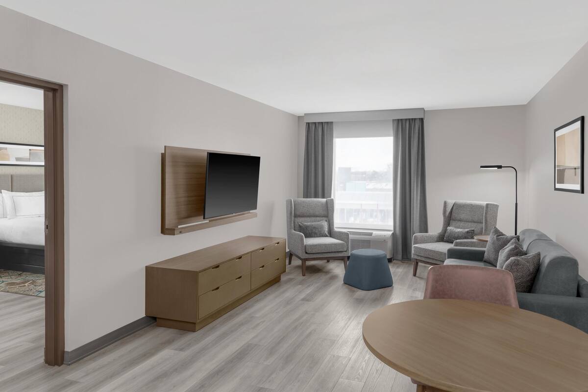 A modern hotel room with a bed, a TV mounted on the wall, a grey armchair, a sofa, a small round table, and a wooden cabinet.
