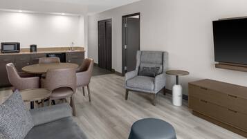 A modern hotel room with a dining area, a gray armchair, a sofa, and a TV mounted on the wall.