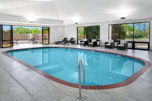 Una piscina interior moderna con agua azul cristalina, rodeada de sillones de descanso y un patio con vistas a árboles y una valla.