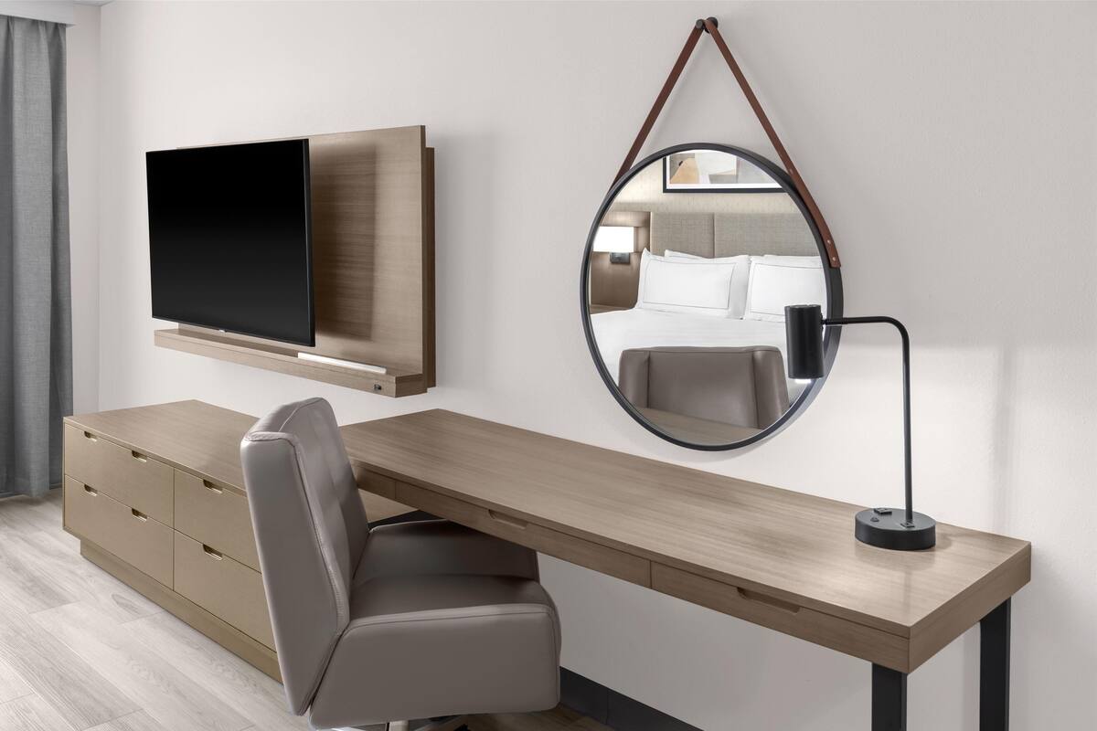 A modern hotel room with a wooden desk, a flat-screen TV, a round mirror, and a chair.