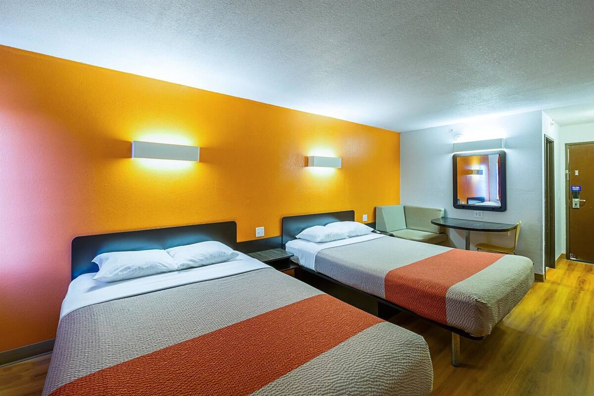 A hotel room with two beds, a small table, a chair, and a mirror on the wall.