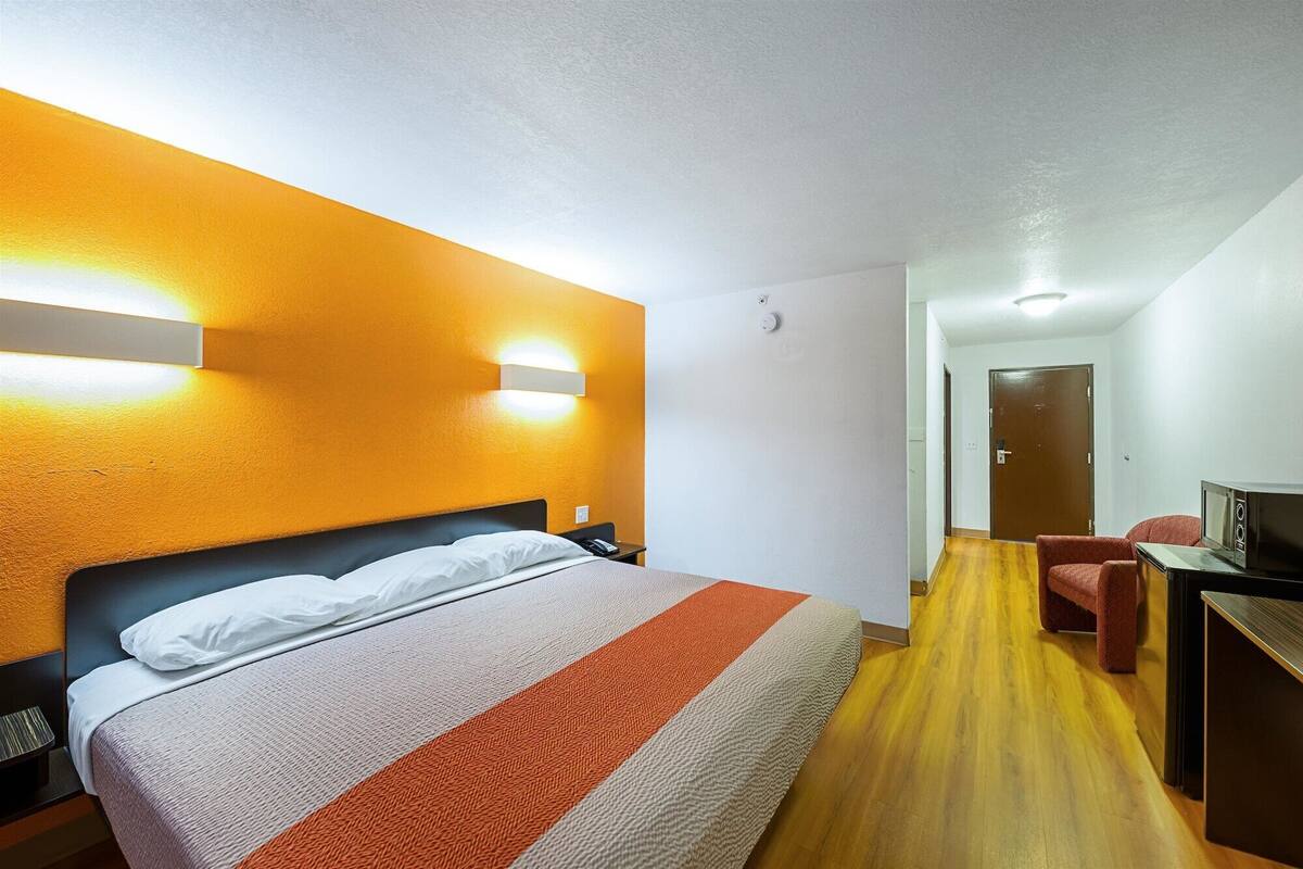 A hotel room with a bed, a desk with a computer, and a door in the background.