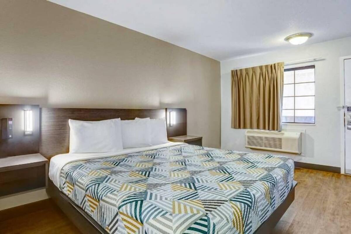 A hotel room with a bed, nightstands, a window with curtains, and an air conditioning unit.