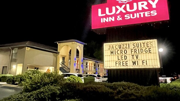 Segnale pubblicitario per 'Luxury Inn & Suites' con servizi tra cui suite con vasca idromassaggio, mini-frigorifero, TV LED, WiFi gratuito e un grande cartello illuminato.