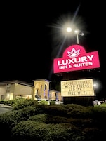Un letrero indica 'Luxury Inn & Suites' y enumera servicios como suites con jacuzzi, refrigerador minibar, TV LED, WiFi gratuito y un gran letrero iluminado.