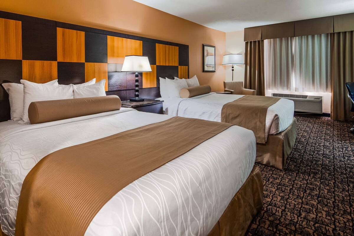 A hotel room with two beds, a nightstand, a lamp, a chair, and a window with curtains.