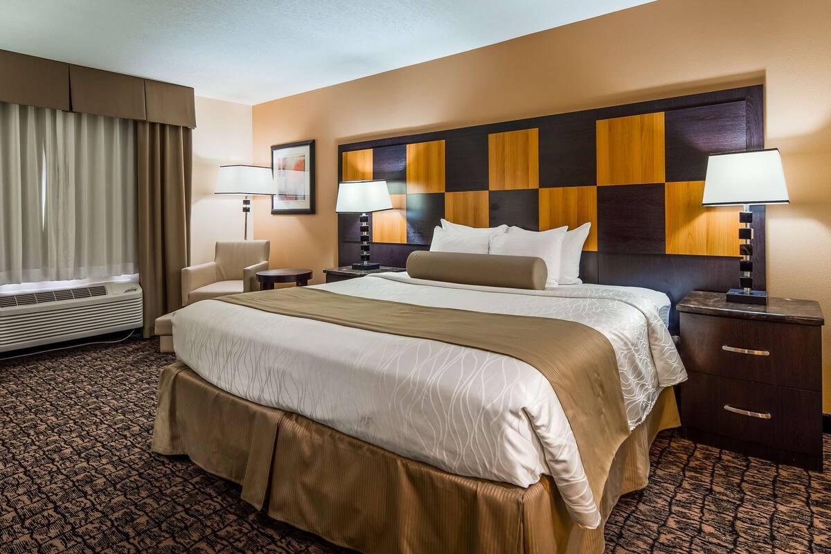 A hotel room with a large bed, two bedside tables, a desk, and a chair. There is a window with curtains, a wall-mounted air conditioner, and a headboard with a checkered pattern.