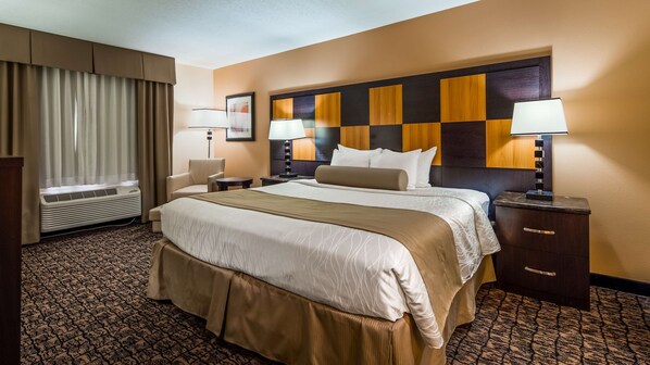 A hotel room with a large bed, two bedside tables, a desk, and a chair. There is a window with curtains, a wall-mounted air conditioner, and a headboard with a checkered pattern.
