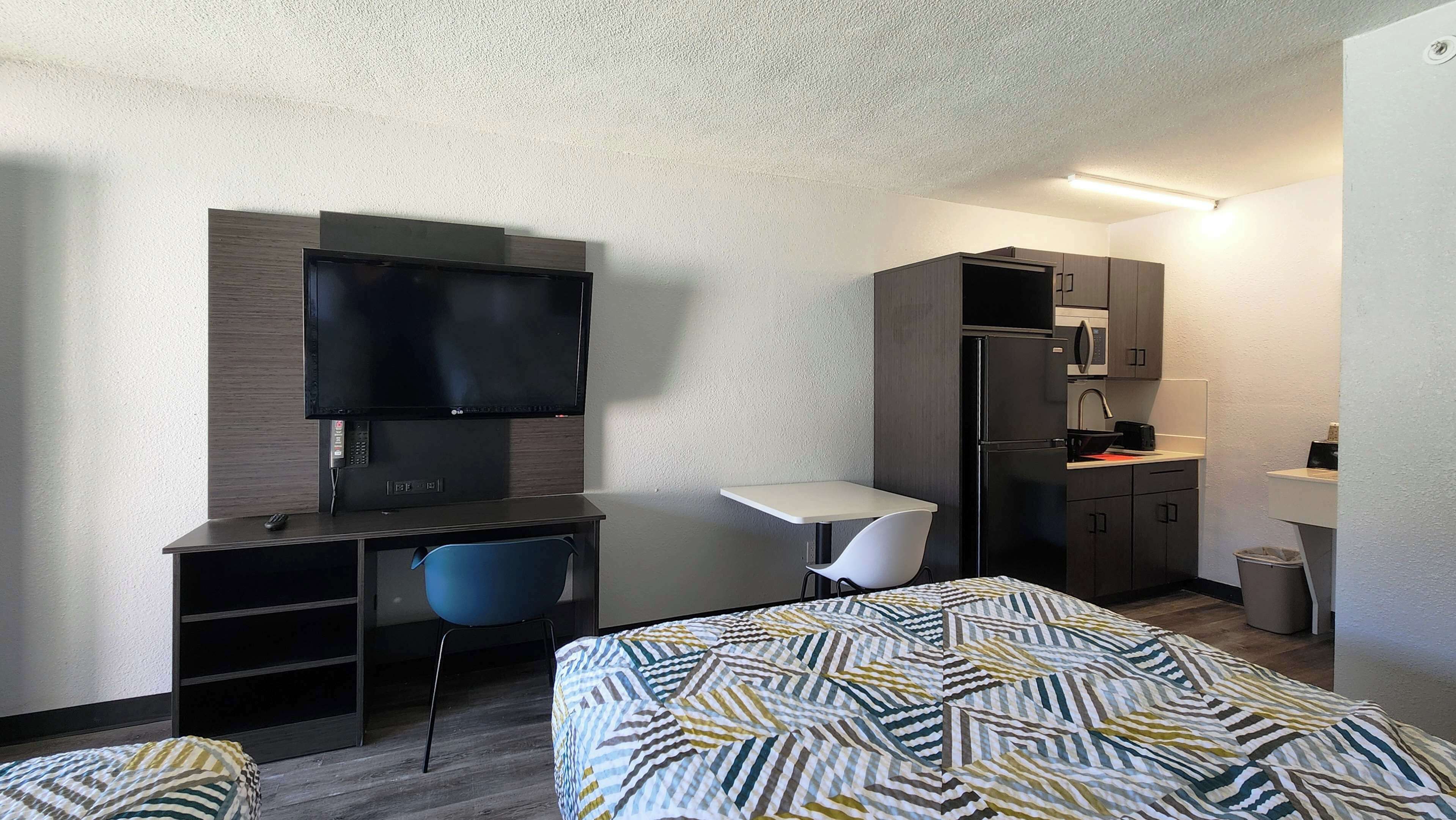 Habitación de hotel con cama, escritorio y silla, televisor, refrigerador, microondas y una pequeña kitchenette.