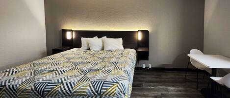A modern hotel room with a large bed, a bedside table, a chair, and a wall-mounted light fixture.
