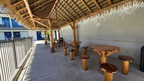 Area salotto esterna con tavoli e sedie di legno, tetto in paglia e vista su un edificio con porte blu.