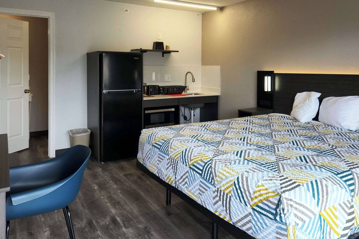 A hotel room with a bed, a desk with a refrigerator, a chair, and a nightstand.