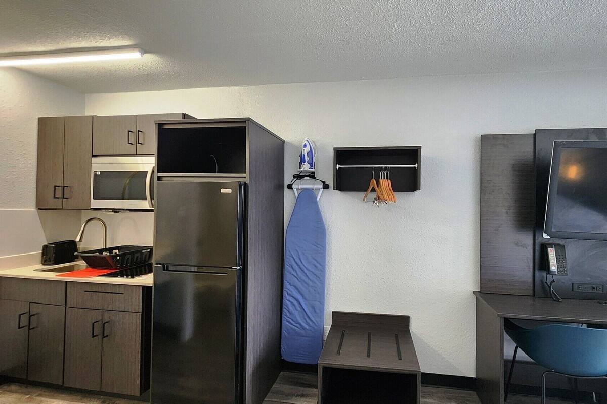 A compact kitchenette with a refrigerator, microwave, and sink. There is a small ironing board and a TV on a stand.