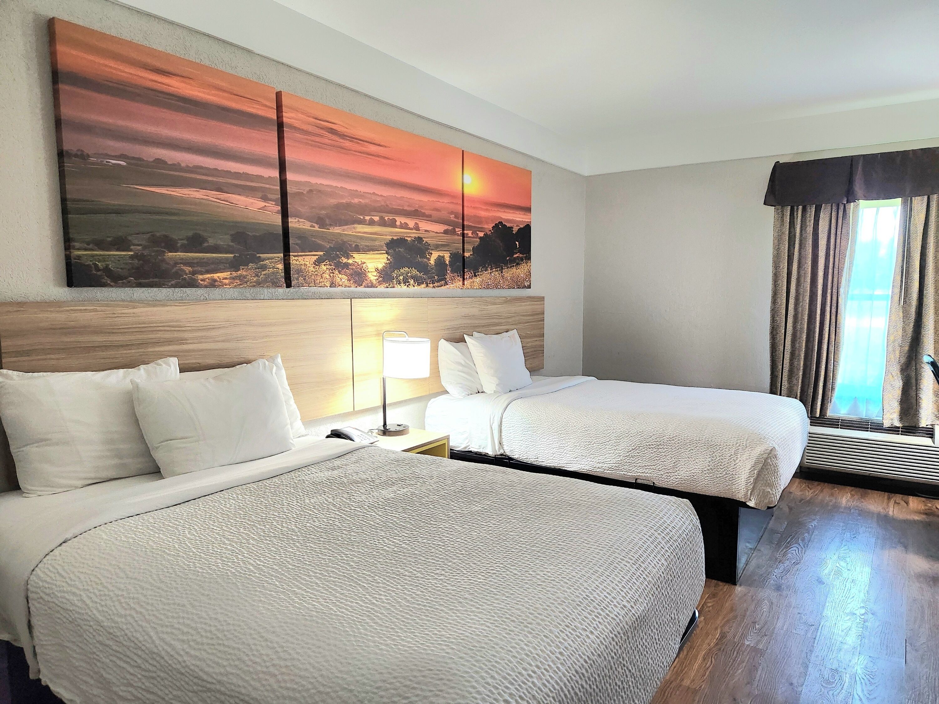 Hotelkamer met twee bedden, een groot schilderij van een zonsondergang, een nachtkastje met een lamp en een raam met gordijnen.