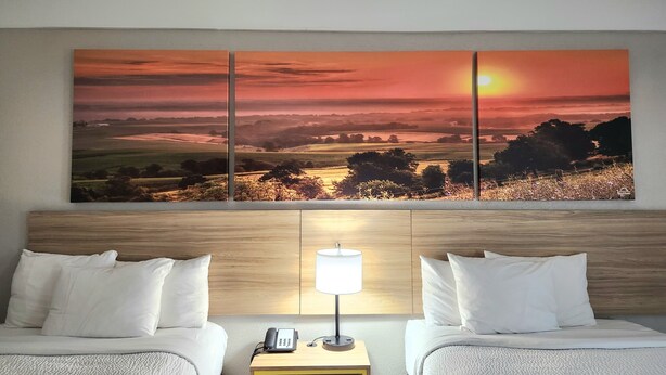 Een hotelkamer met twee bedden, een nachtkastje met een lamp, en een groot landschap schilderij boven de bedden.