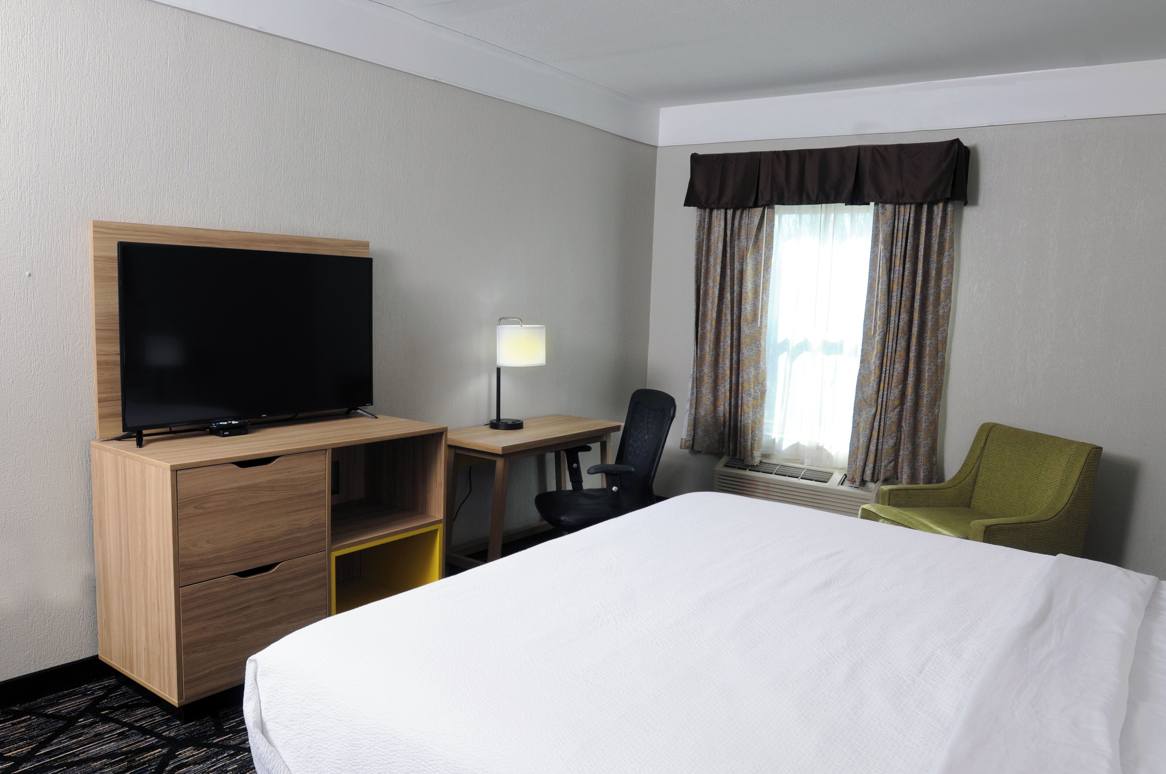 A hotel room with a bed, a TV on a wooden cabinet, a desk with a chair, and a window with curtains.