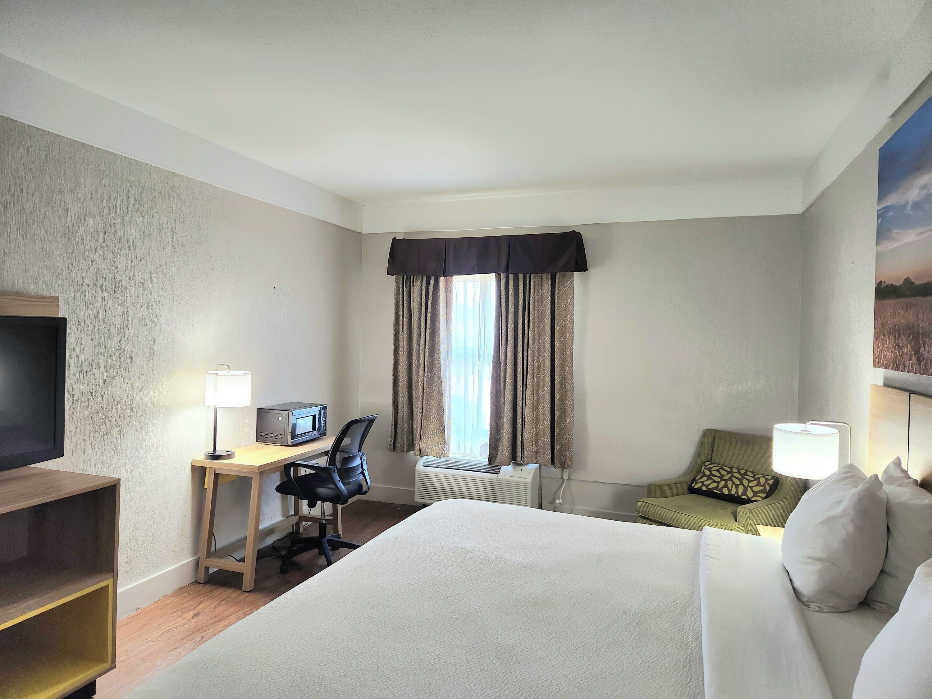 Een hotelkamer met een groot bed, een bureau met computer en magnetron, een televisie, een stoel en een raam met gordijnen.