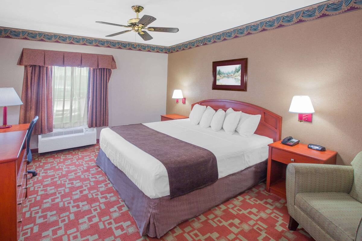 A hotel room with a bed, two bedside tables with lamps, a chair, a desk with a lamp, a ceiling fan, and a window with curtains.