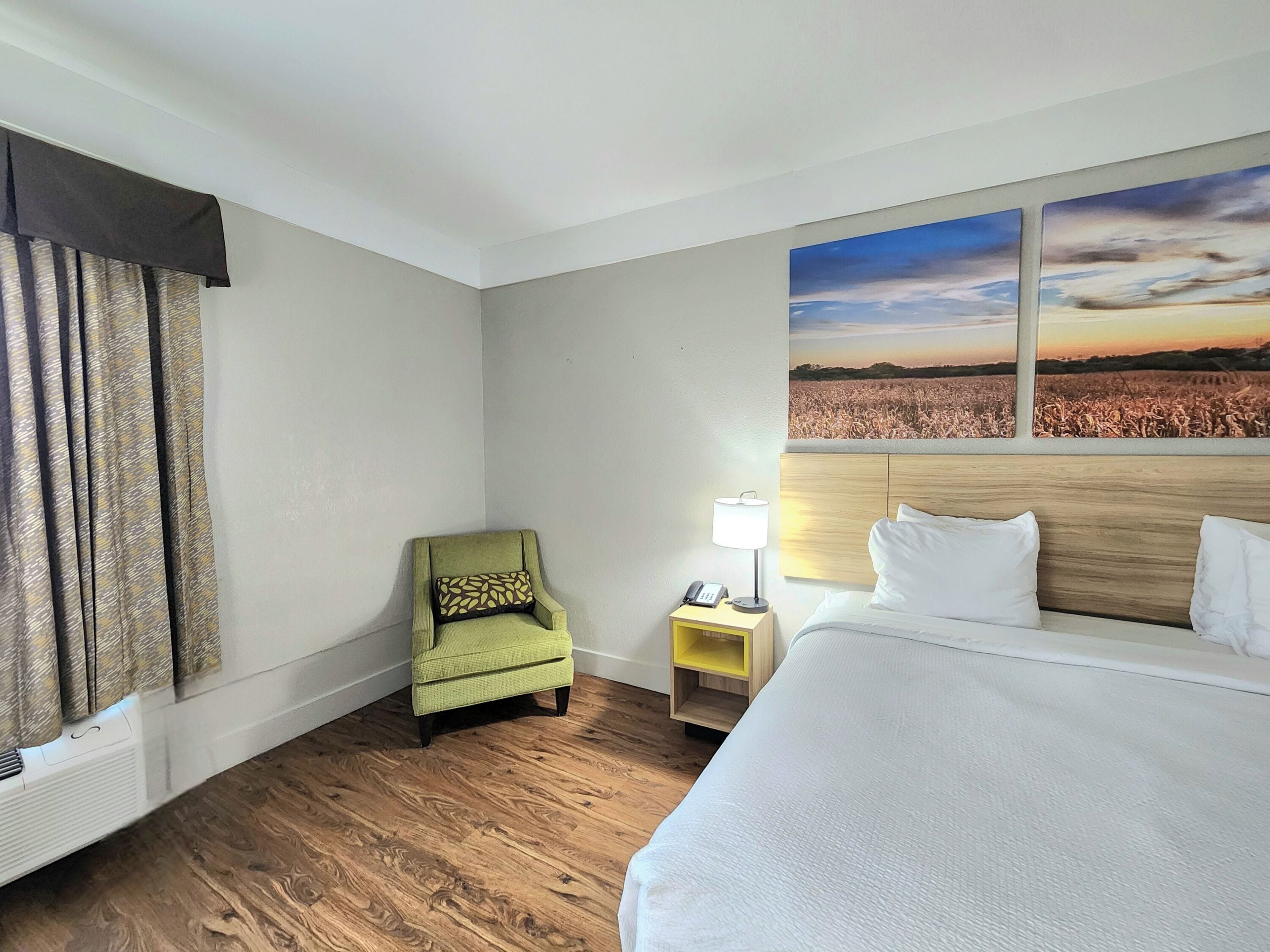 Ein Hotelzimmer mit einem großen Bett, einem Sessel, einem Nachttisch mit Lampe und einem Wandbild, das einen Sonnenuntergang über einem Feld zeigt.