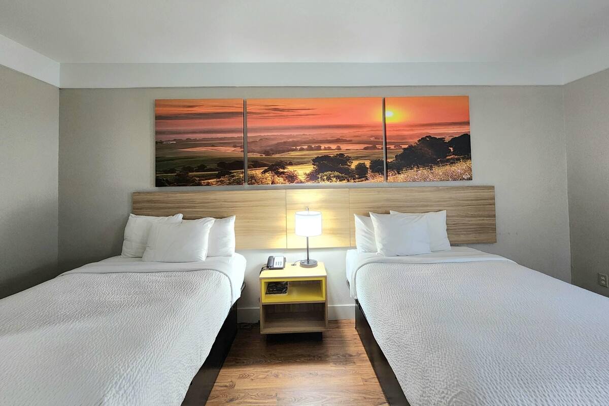 A hotel room with two beds, a wooden headboard, a nightstand with a lamp, and a large landscape painting above the beds.