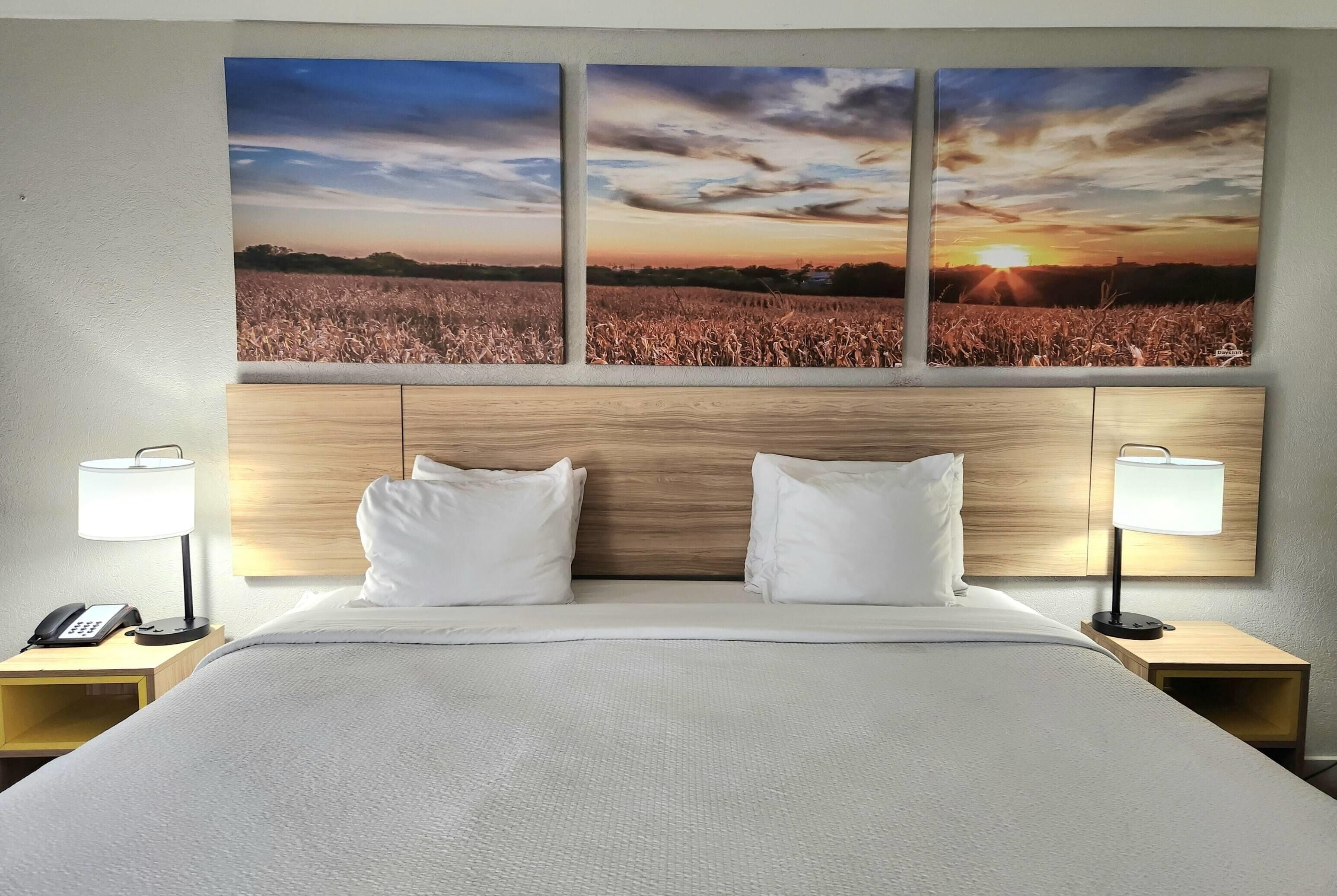 A hotel room with a large bed, two bedside tables with lamps, a phone on one table, and a wall-mounted painting of a sunset over a field.