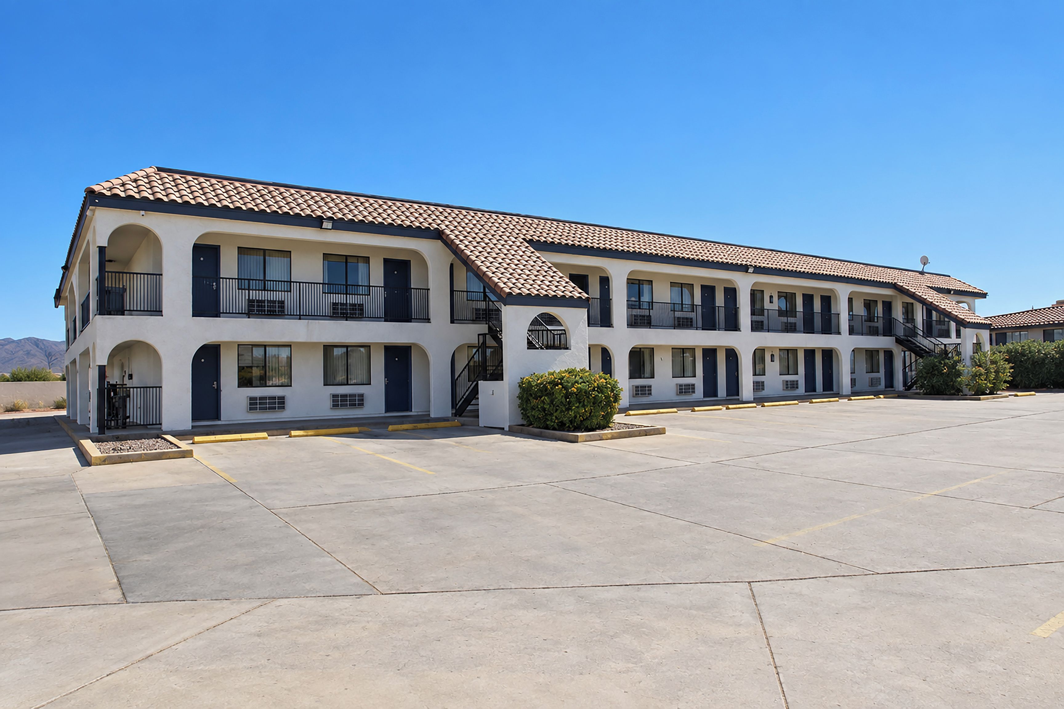 A two-story building with a flat roof, multiple balconies, and a parking lot in front.