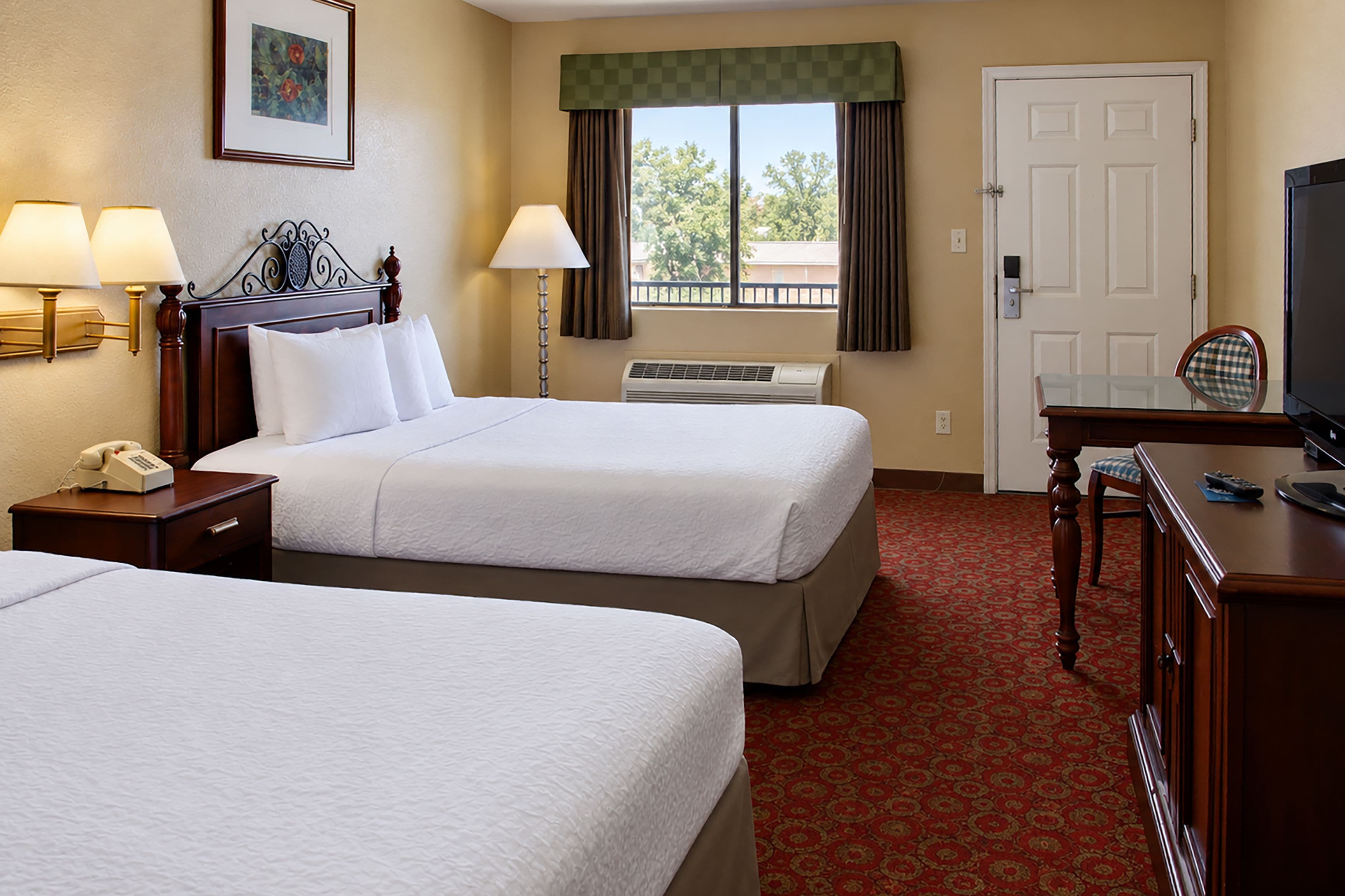 A hotel room with two beds, a desk with a television, a telephone, and a window with curtains.