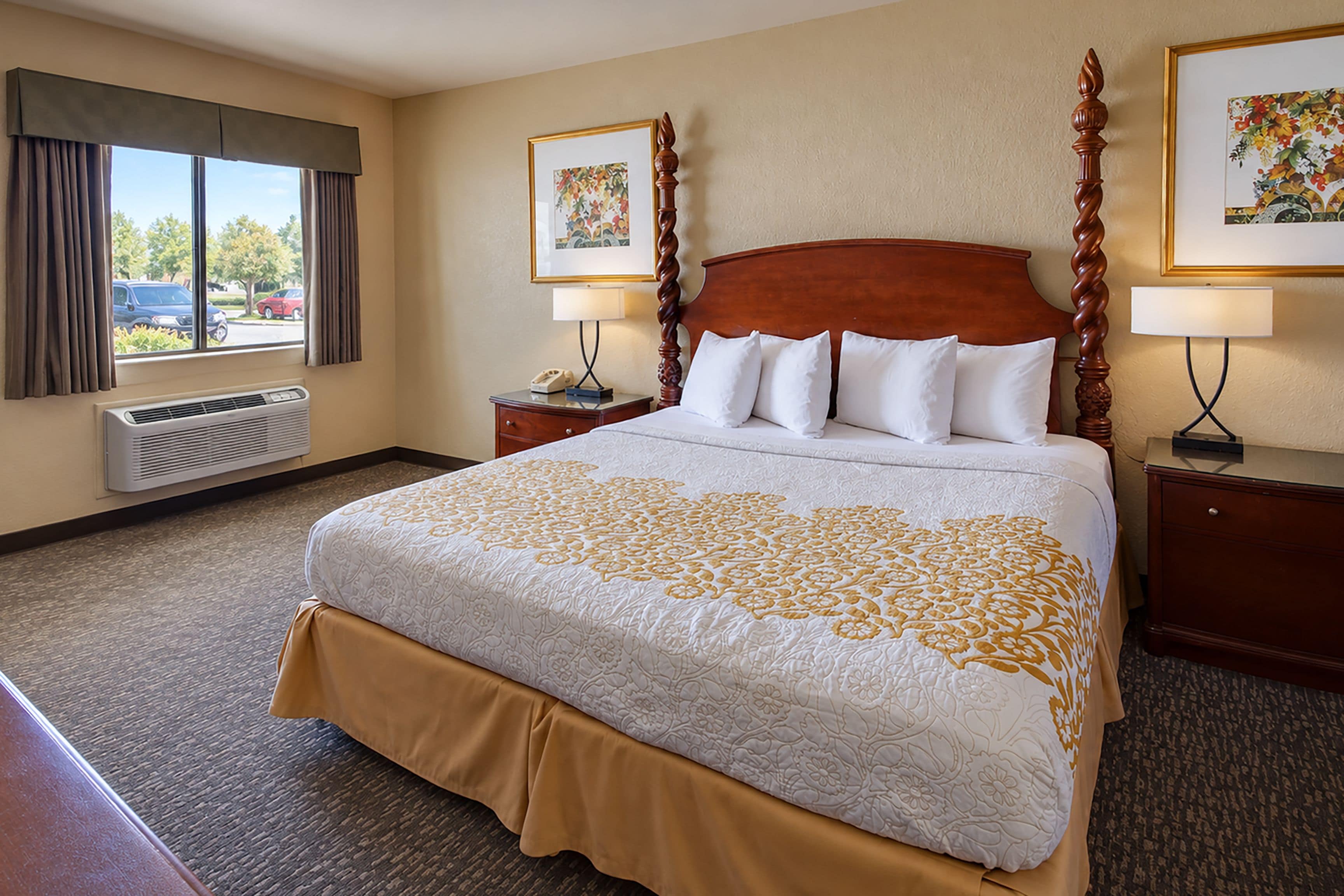 A hotel room with a large bed, two bedside tables, a window with curtains, an air conditioning unit, and two framed pictures on the wall.