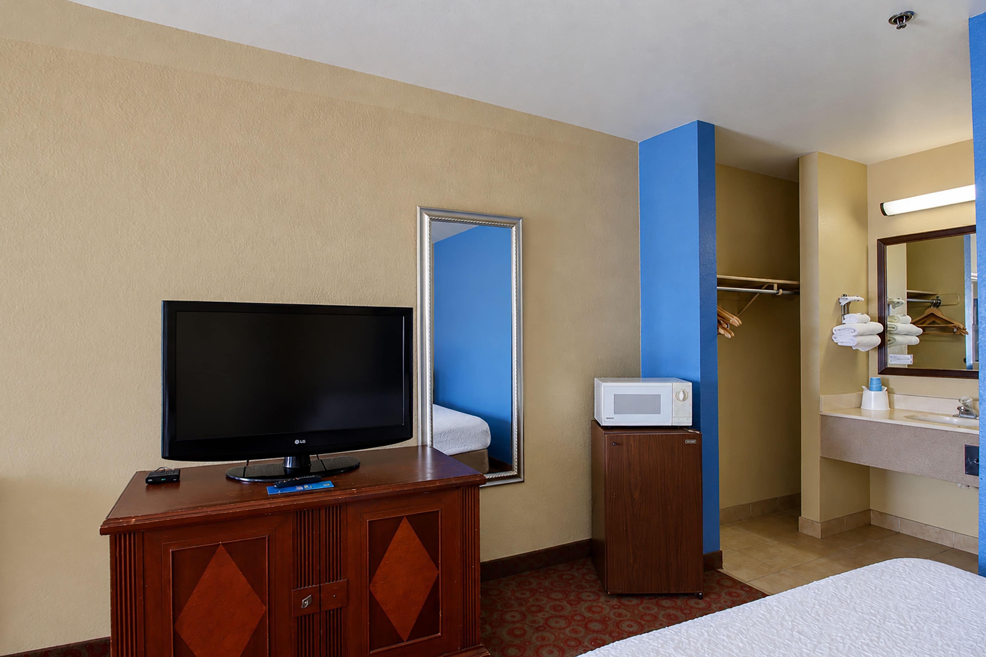 A hotel room with a television on a wooden cabinet, a microwave, a bathroom with a sink and mirror, and a bed with a white comforter.