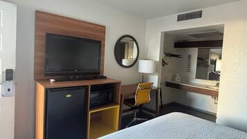 A hotel room with a bed, a television, a desk with a chair, a mirror, and a small refrigerator.