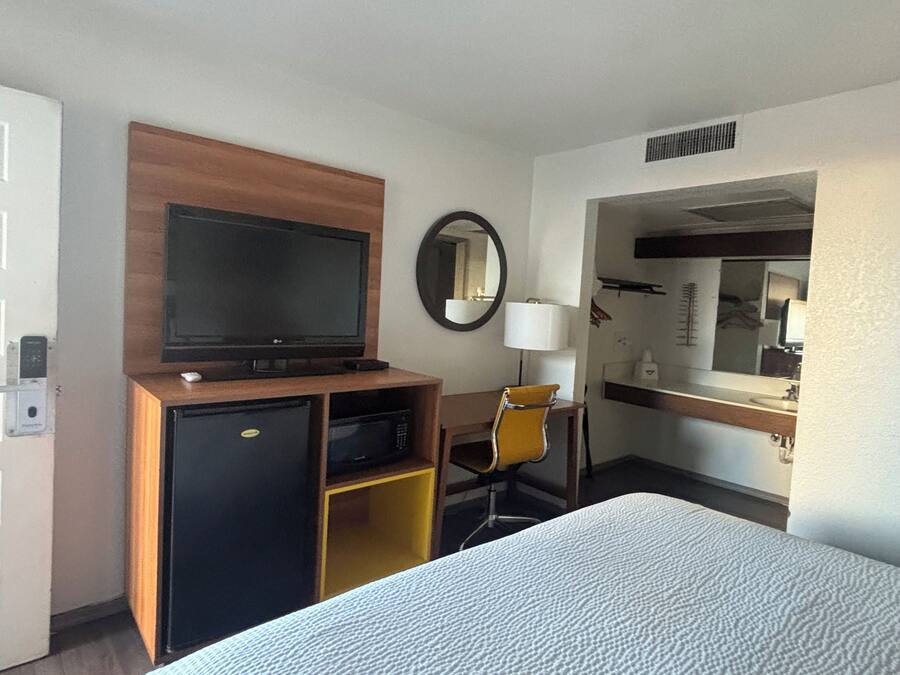 A hotel room with a bed, a television, a desk with a chair, a mirror, and a small refrigerator.