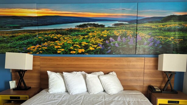 Una habitación de hotel con una cama grande, ropa de cama blanca y un cabecero de madera. Hay dos mesitas de noche con lámparas y un atril con un teléfono.