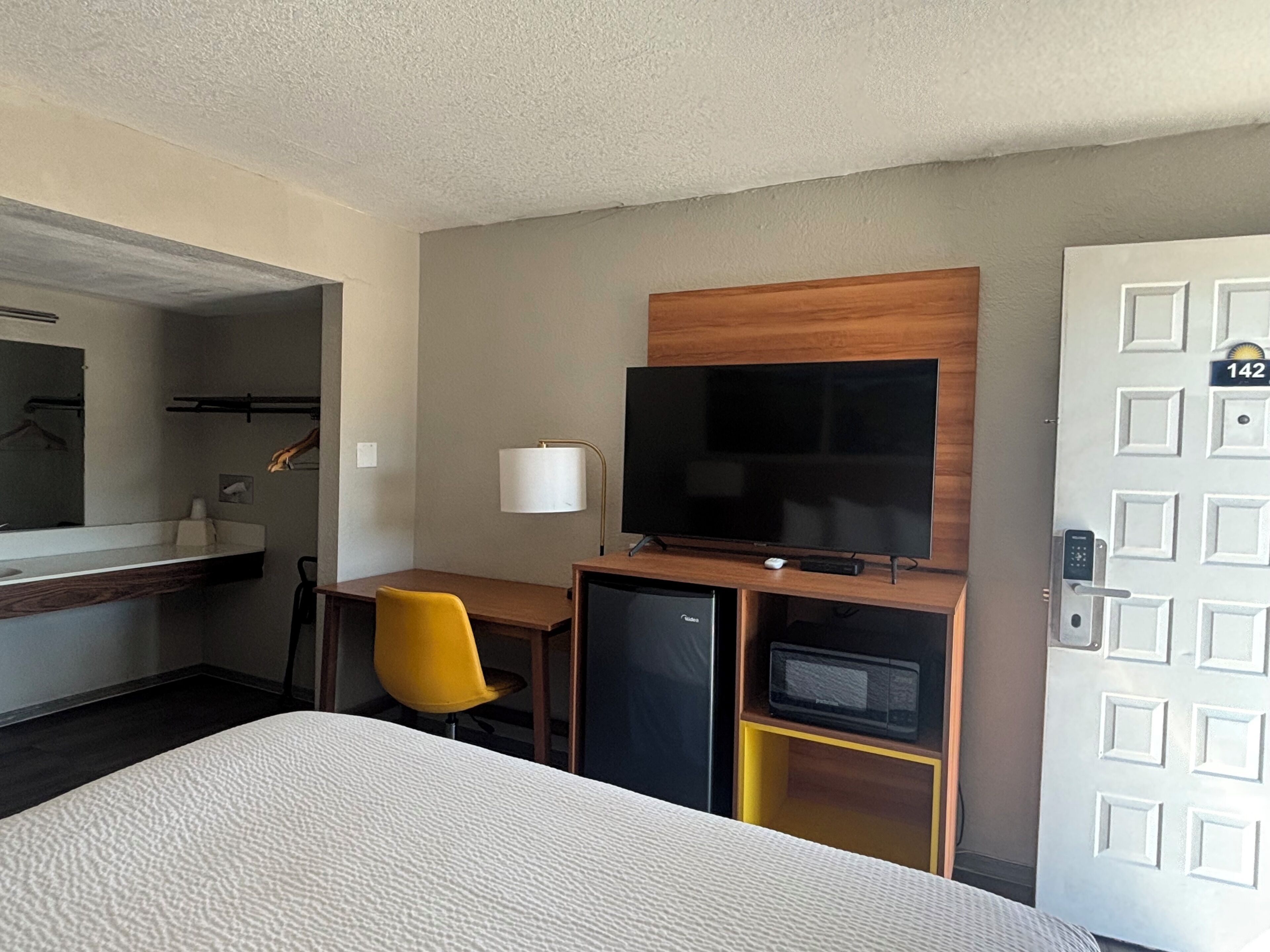 Habitación de hotel con cama, escritorio, silla, televisor, minibar y puerta con placa de número.