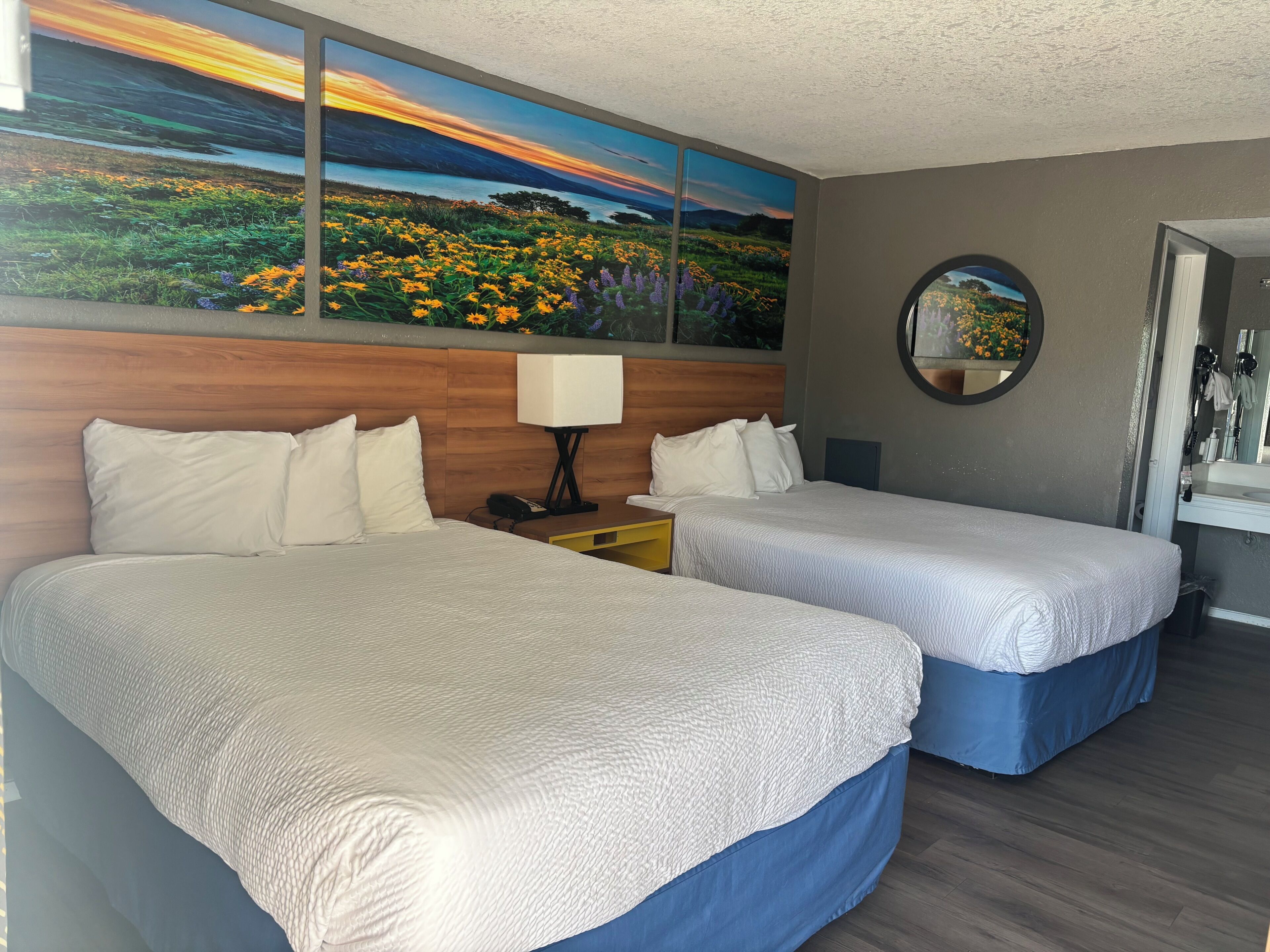 Habitación de hotel con dos camas, una mesita de noche, una lámpara, un cuadro de un paisaje y un espejo.