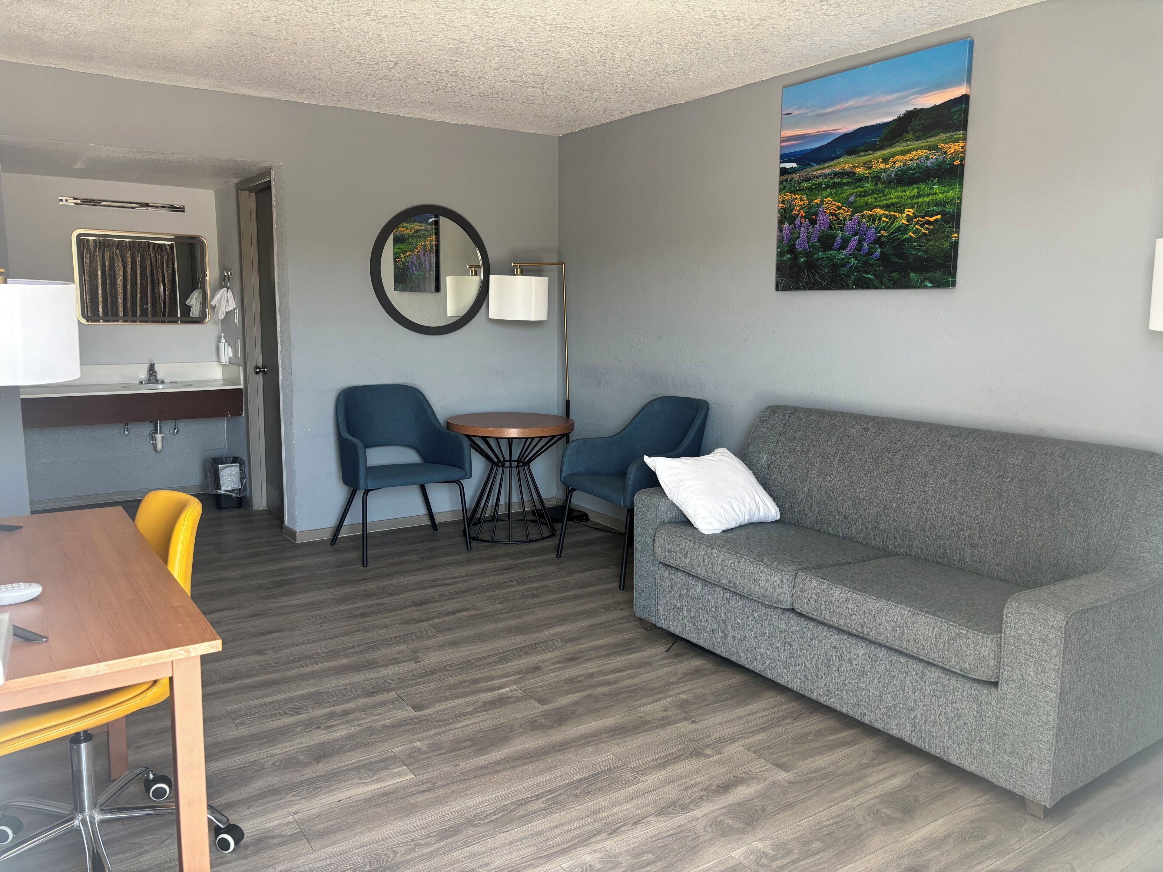 Ein modernes Wohnzimmer mit einem grauen Sofa, zwei blauen Sesseln, einem Holzesstisch und einem kleinen runden Tisch mit einer Lampe.