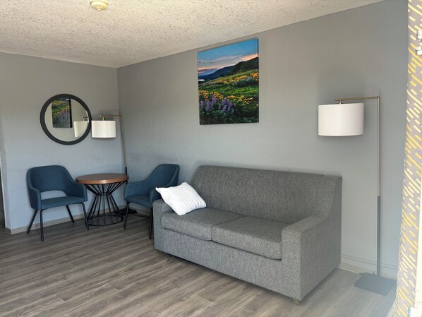 A modern living room with a grey sofa, two blue armchairs, a round coffee table, a wall-mounted mirror, and a landscape painting.
