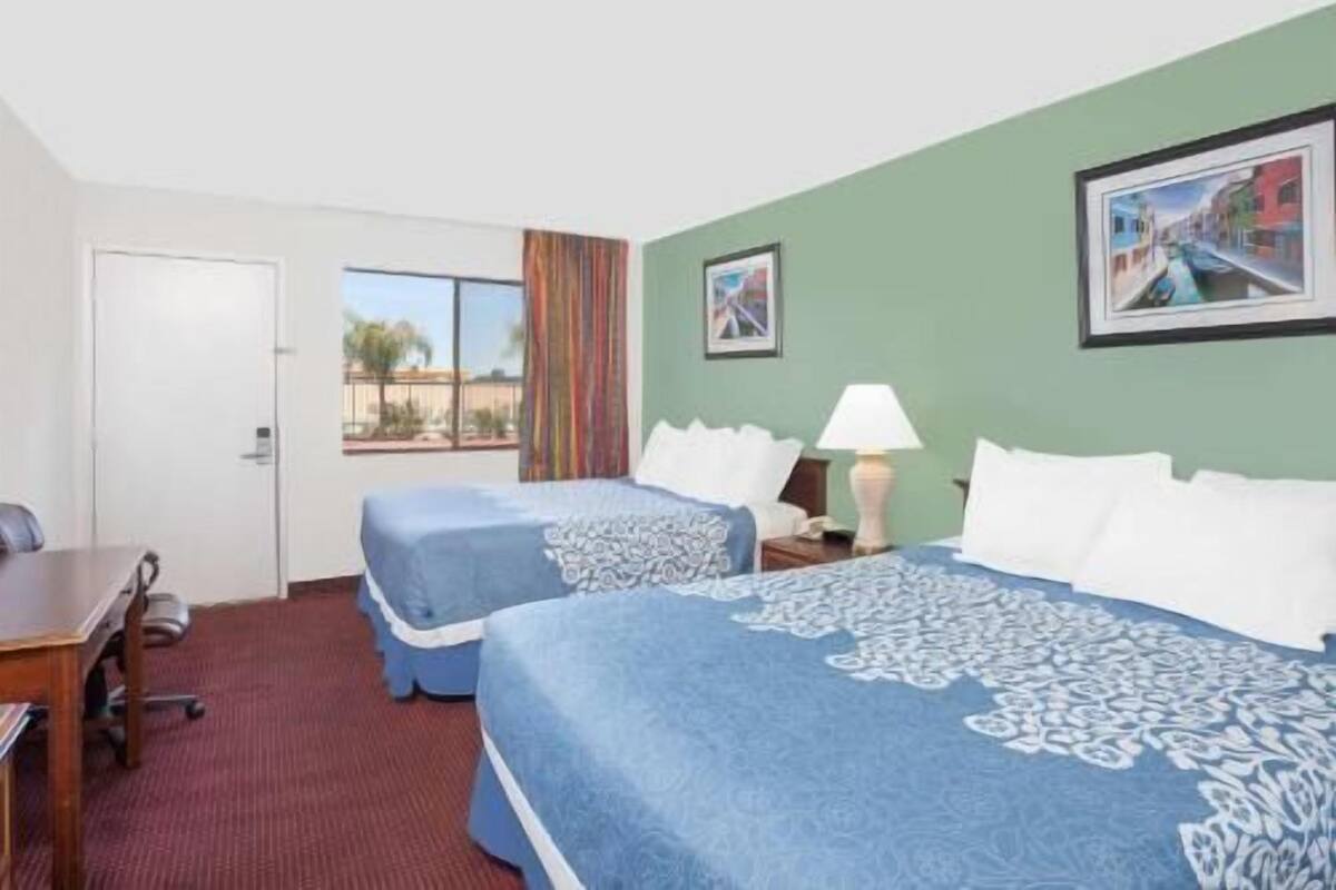 A hotel room with two beds, a desk, a chair, a window with a view, and a framed picture on the wall.