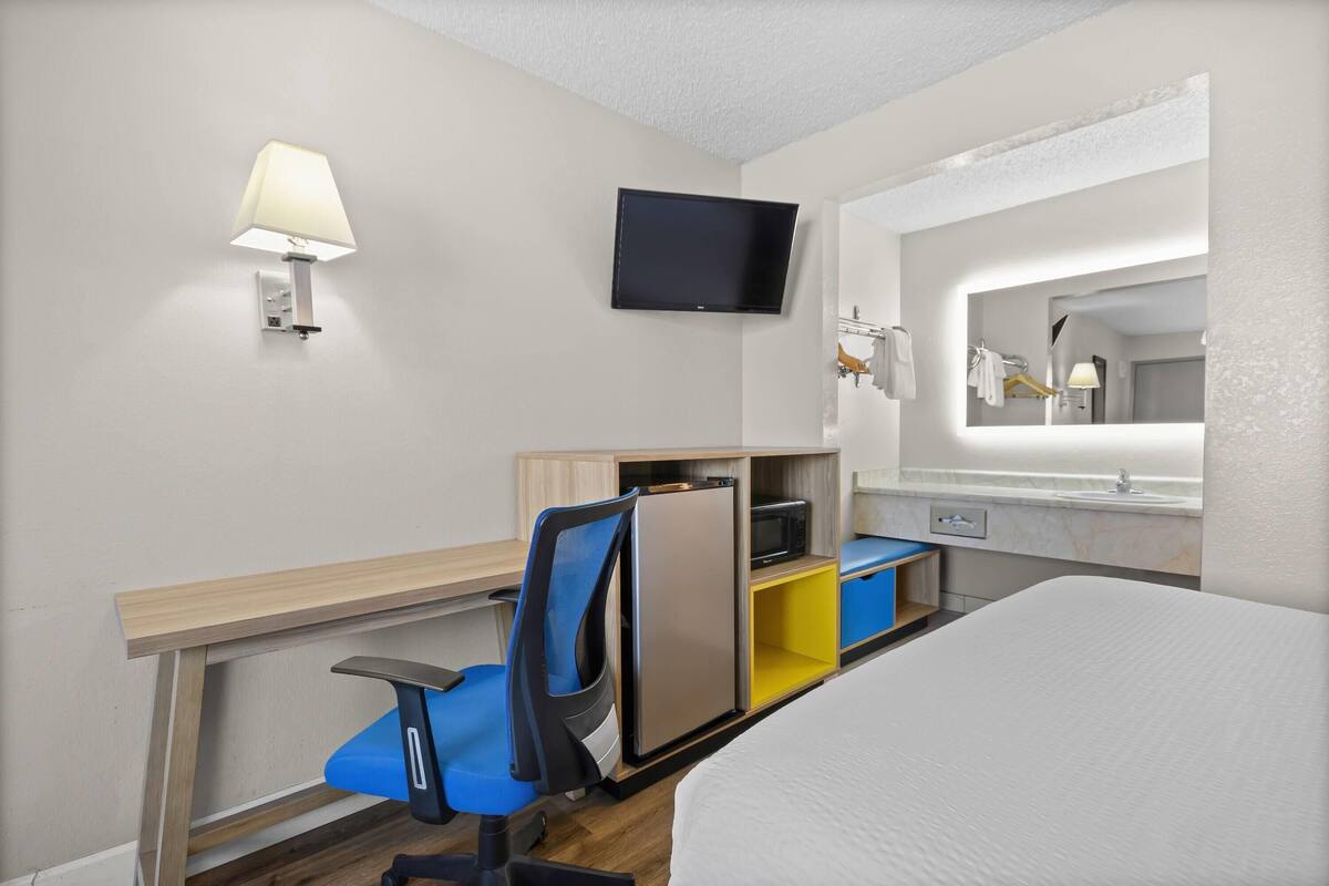 A hotel room with a bed, a desk with a chair, a TV mounted on the wall, a mirror, and a small shelf with a lamp.