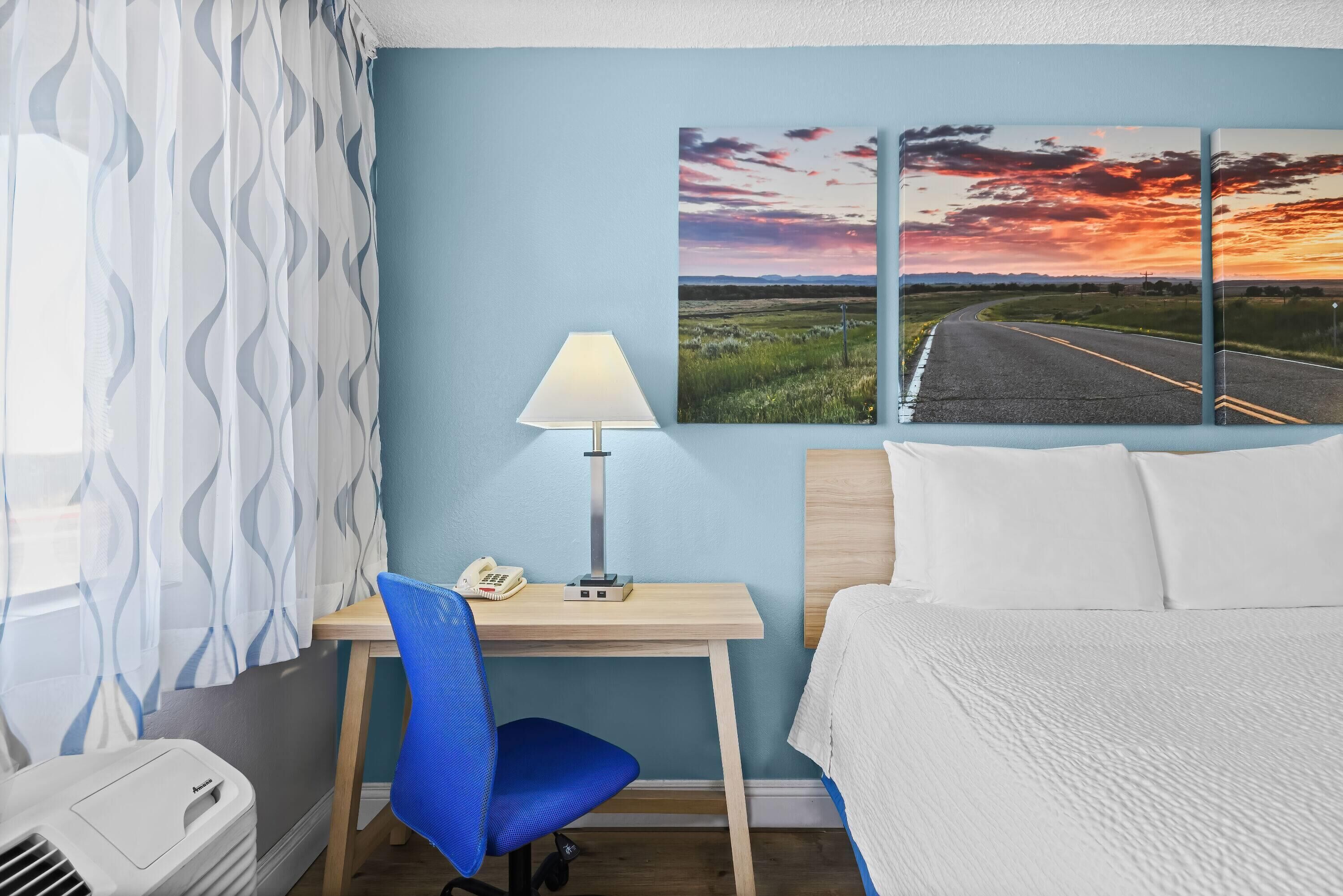 A hotel room with a bed, a desk with a lamp, a chair, a phone, and a heater.