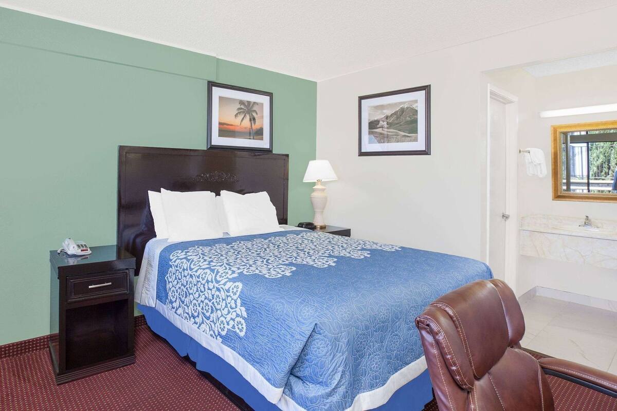 A hotel room with a bed, a nightstand, a chair, a lamp, and a framed picture on the wall.